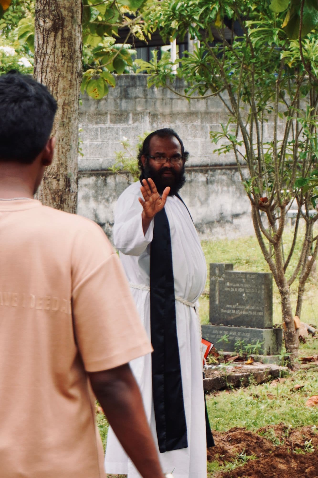 The priest in white robe and black stole, hand raised mid-blessing among the graves.